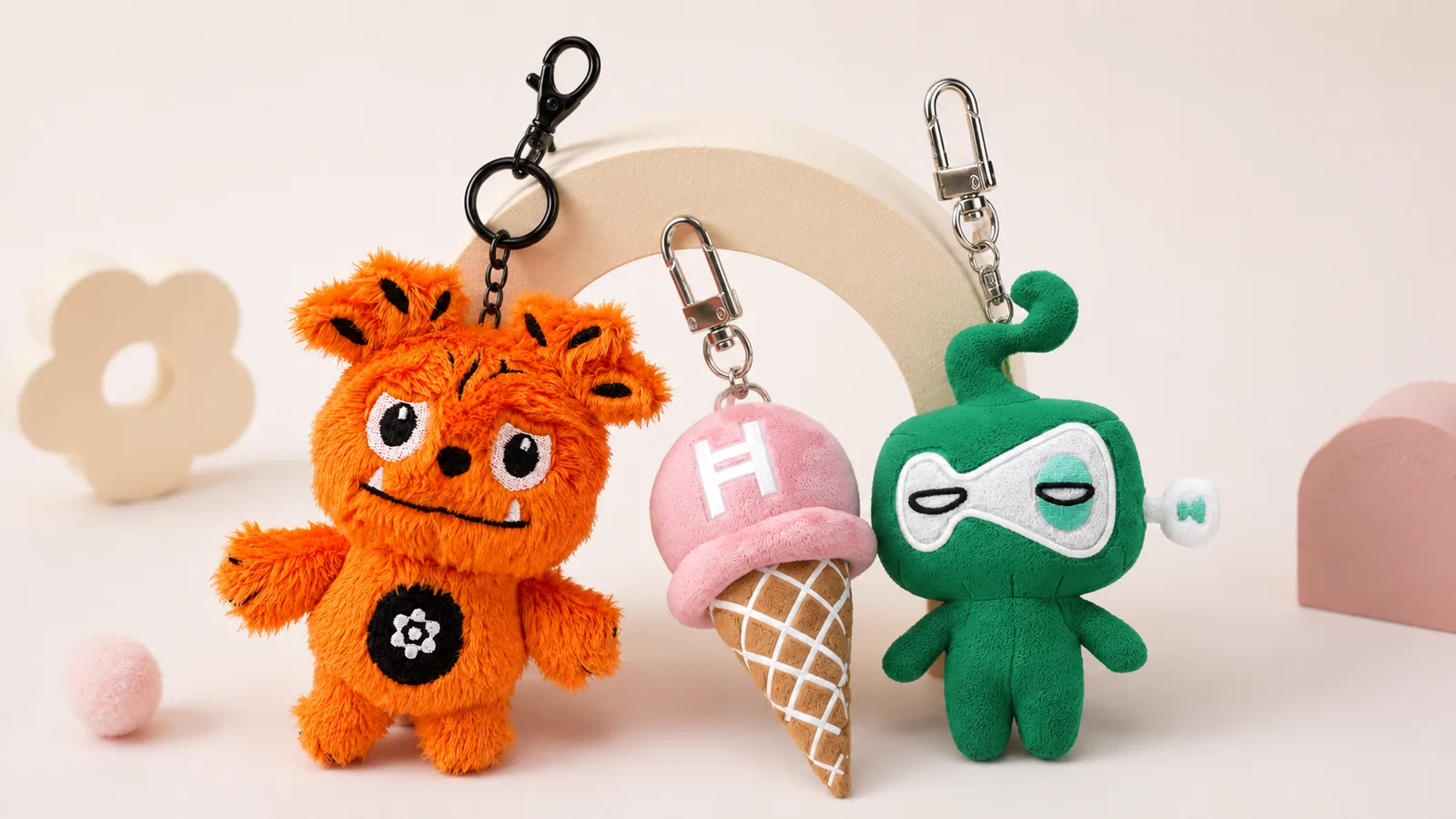 Three character plush keyrings, an orange monster, a pink ice cream, and a green robot, lined up on a pastel background